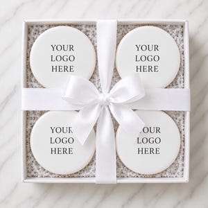 May include: A square white gift box containing four round white coasters, each printed with the black text YOUR LOGO HERE. The box is tied with a white satin ribbon featuring a central bow, and the coasters rest on a bed of white shredded paper.