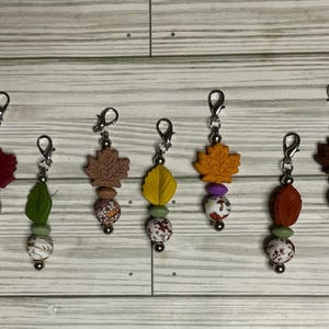 Op de afbeelding: Zeven decoratieve bedels met herfstachtige siliconen bladvormige hangers in rood, bruin, geel en oranje. Elke bedel is voorzien van een zilverkleurige karabijnsluiting, kleine ronde tussenkralen en siliconen kralen met bloemen- of dierenprints.