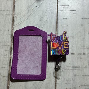 Peut inclure: Un porte-badge vertical violet accompagné d'un enrouleur de badge assorti avec un motif graphique coloré. L'enrouleur affiche le texte "teach love inspire" avec une icône de pomme rouge et des motifs de crayons sur un fond violet.