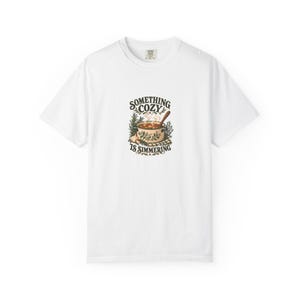 May include: A white short-sleeve t-shirt featuring a central graphic of a cooking pot with steam, surrounded by herbs and garlic. The text "SOMETHING COZY IS SIMMERING" is printed in dark brown lettering above and below the illustration.