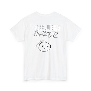 Könnte beinhalten: Ein weißes kurzärmeliges T-Shirt mit schwarzem Text, der TROUBLE MAKER in einer Mischung aus Block- und Schreibschrift liest. Unter dem Text befindet sich ein kleines, handgezeichnetes kreisförmiges Gesicht mit geschlossenen Augen und einem Lächeln, begleitet von dem Text Chaos with good intentions.