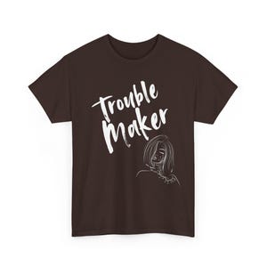 Könnte beinhalten: Ein dunkelbraunes T-Shirt mit kurzen Ärmeln und dem weißen Schriftzug Trouble Maker in Pinselstrich-Optik. Unter dem Text befindet sich eine weiße Strichzeichnung einer Person mit kurzem Haar und einem Blumenmuster an Hals und Schulter.