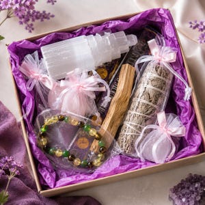 May include: A gift box lined with purple tissue paper containing a white selenite tower, a bundle of dried sage wrapped in string, a wooden stick, a beaded bracelet with green and yellow stones, and several small pink organza drawstring bags.