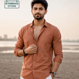 May include: A long-sleeved, button-down shirt in a solid rust-orange color with a classic collar, paired with white trousers. The shirt features a textured fabric, a front button placket, and sleeves rolled up to the elbows, worn with a dark wristwatch.