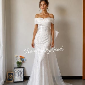 May include: A floor-length white off-the-shoulder gown featuring a draped bodice, a fitted silhouette, and a sheer overlay with subtle vertical embellishments. The dress includes a large bow detail at the back and a long, flowing train. Text reads Rainbowsky Goods.