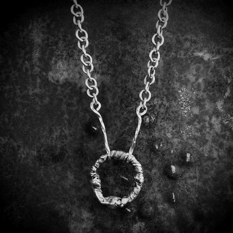 May include: A silver-toned necklace featuring a circular pendant with a textured, uneven surface. The pendant is suspended from a chain composed of interconnected round links that transition into two twisted wire segments at the attachment point.