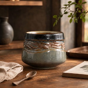 May include: A ceramic bowl with a wide, rounded base and a tapered rim. The exterior features a textured, wavy band in earthy brown tones, contrasted by a smooth, muted sage green lower section and a dark, glossy black glaze around the top rim.