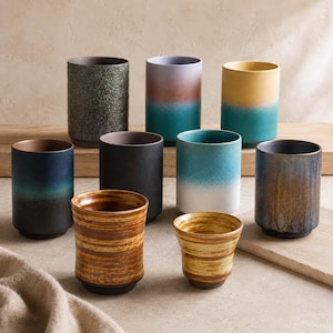 May include: A collection of nine ceramic cups in various finishes and colors, including textured dark gray, gradient blue-to-teal, yellow-to-teal, solid matte black, white-to-teal, brown-to-blue, and two cups with horizontal brown and yellow stripes.