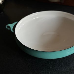 May include: A round, shallow teal enameled cast iron pan with a white interior. The pan has two teal handles and a black rim. This cookware is suitable for cooking and serving.