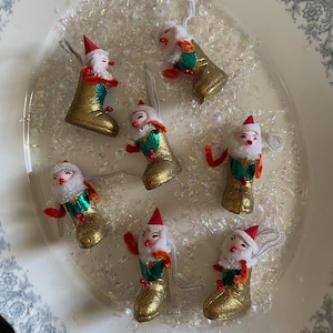 May include: A collection of vintage Christmas ornaments shaped like Santa Claus figures in gold boots. Each ornament has a red hat, white beard, and red and green accents. They are arranged on a decorative plate with glitter.