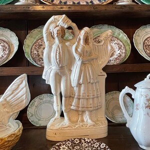 May include: A white ceramic figurine of a couple in traditional dress. The man is holding a hat and the woman is holding a harp. The figurine is on a white base with a floral design.