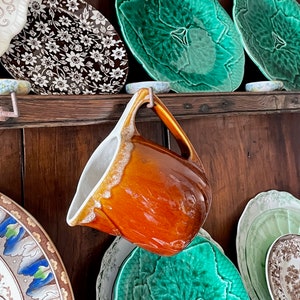 May include: A collection of vintage ceramic plates and a brown glazed pitcher hanging on a wooden shelf. The plates feature floral and leaf patterns in shades of green, white, and brown.