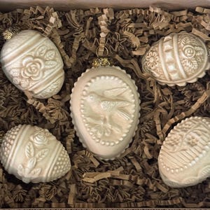 May include: Five off-white Easter egg ornaments with gold-colored caps, nestled in brown paper. The eggs feature embossed floral and bird designs. The ornaments are in a cardboard box.