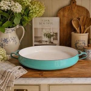 May include: A round, shallow, turquoise enameled cast iron braiser pan with two side handles and a smooth white interior. The pan is positioned on a wooden surface in front of a cookbook titled Cook Beautiful, a white pitcher, and a ceramic crock.