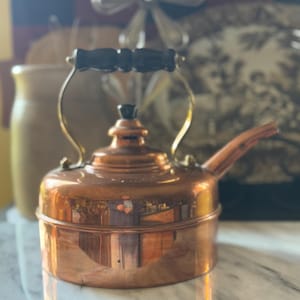 May include: A copper tea kettle with a black handle and spout. The kettle has a shiny, reflective surface and a brass handle. The design is vintage, with a classic shape and a small, decorative knob on the lid.