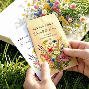 May include: Two rectangular paper seed packets featuring the printed text LET LOVE GROW, Barnett & Amelia, 05.05.2026, and WILDFLOWER SEEDS. The packets are decorated with a colourful illustration of a wildflower bouquet including poppies and daisies.