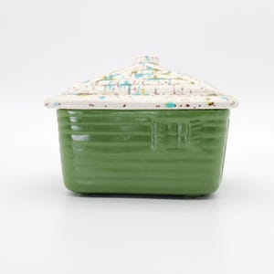 May include: A ceramic container shaped like a small house with a green base featuring horizontal siding and a window. The removable lid is white with a textured shingle pattern and is decorated with blue, yellow, and brown paint splatters.