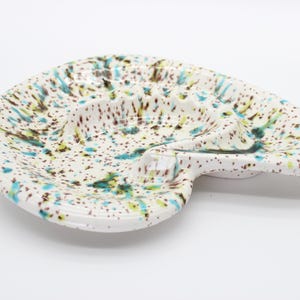 May include: A white ceramic ashtray with an irregular, kidney-like shape featuring a speckled pattern of brown, teal, and yellow paint splatters. The surface has a glossy finish with a central indentation and a small notch on the side.