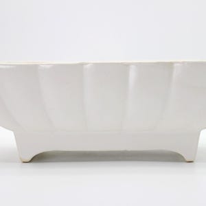 May include: A rectangular white ceramic planter featuring a scalloped exterior design with vertical ridges. The vessel is supported by four short, sturdy feet at the base, providing a raised profile for this functional piece of home decor.