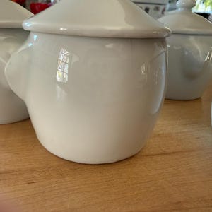 May include: A collection of white ceramic lidded pots with smooth, rounded bodies and small side handles. Each pot features a matching domed lid with a small knob handle on top. The glossy white finish reflects light, highlighting the curved shape.
