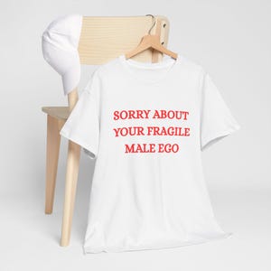 Può includere: Una t-shirt bianca a maniche corte con il testo rosso SORRY ABOUT YOUR FRAGILE MALE EGO stampato sul davanti, esposta su una gruccia di legno accanto a un cappellino da baseball bianco tinta unita. La maglietta ha un girocollo e vestibilità standard.
