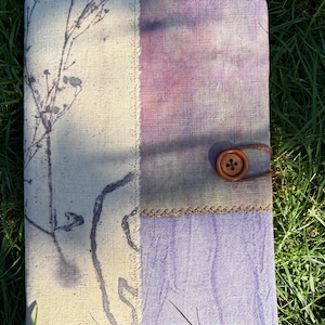 Op de afbeelding: Een rechthoekig, met stof bekleed notitieboek met een crèmekleurig linkerpaneel met een donkere botanische print en een rechterpaneel verdeeld in twee secties van paarse en lavendelkleurige tie-dye stof, vastgezet met een bruine knoop en elastiek.