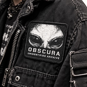 Puede incluir: Un parche bordado negro con un gráfico de cabeza alienígena blanca sobre el texto OBSCURA y UNIDENTIFIED ARCHIVE, cosido en una chaqueta vaquera negra con cremallera metálica, botones plateados y una cadena visible en el fondo.