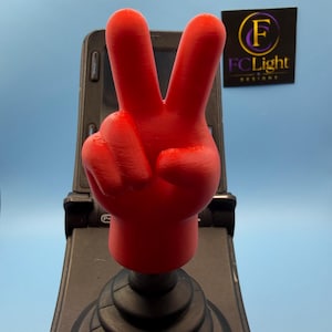 May include: A bright red 3D-printed hand gesture topper shaped like a peace sign, mounted on a black circular joystick base with a ribbed rubber boot. The hand features a smooth finish and is positioned vertically above the black control mechanism.