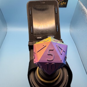 May include: A multi-coloured 20-sided die with a gradient of purple, blue, orange, and yellow, featuring raised black numbers. The die is positioned in front of a black electronic device with a screen and buttons, labelled with the text F Light.