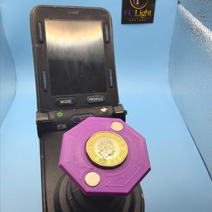 Op de afbeelding: Een paarse achthoekige 3D-geprinte joystick-opzetstuk met een goudkleurige Britse munt van één pond in het midden, gemonteerd op een zwarte elektronische rolstoelbesturing met een scherm dat de knoppen Mode en Profile weergeeft.