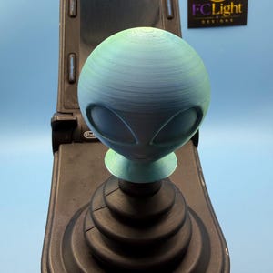 Op de afbeelding: Een 3D-geprinte versnellingspookknop in de vorm van een alienhoofd met een kleurverloop van blauw en groen en zichtbare horizontale laaglijnen. De knop is gemonteerd op een zwarte joystickbasis met een vierkant scherm en een logo met de tekst FCLight DESIGNS.