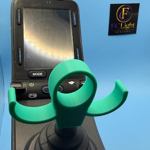 May include: A bright green, 3D-printed joystick handle attachment with a circular top loop and two curved side supports. The accessory is mounted onto a black joystick base featuring a digital display screen with buttons labelled MODE and P-M.