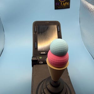 以下が含まれることがあります： ライトブルーの上段とピンクの下段の2段アイスクリームコーンの形をしたジョイスティックコントローラー用トッパー。黒いベースに取り付けられています。コントローラーには電源ボタンとモードボタン付きの画面があり、FCLightのロゴが表示されています。