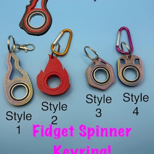 May include: Five fidget spinner keyrings labelled Style 1 through 5. Style 1 is iridescent, Style 2 is red, Style 3 is purple-toned, Style 4 is multi-coloured, and Style 5 is black and red. Each features a central metal bearing and a metal clip.