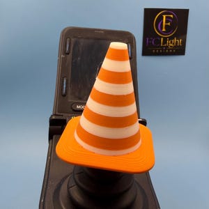 May include: A small, orange and white striped traffic cone topper sits atop a black joystick controller. The cone features horizontal bands and a square base. In the background, a black square sign displays the text F FCLight DESIGNS.