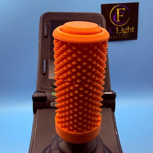 Peut inclure: Une poignée de joystick cylindrique orange vif présentant une surface texturée avec de nombreuses petites pointes. La poignée est fixée sur une base noire avec un soufflet en caoutchouc circulaire à gradins devant un panneau de commande noir.