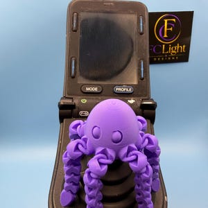 Op de afbeelding: Een 3D-geprint paars octopusfiguur met beweegbare tentakels en een gladde ronde kop, geplaatst op een zwart elektronisch apparaat met een scherm en knoppen met de tekst MODE en PROFILE, naast een bordje met FCLight DESIGNS.