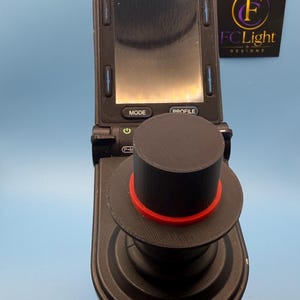 Puede incluir: Un controlador de joystick negro con una parte superior cilíndrica y un anillo rojo. El dispositivo incluye una pantalla digital con botones etiquetados como MODE y PROFILE, y un icono de encendido. Un letrero negro con el texto FCLight DESIGNS es visible.