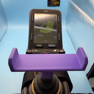 May include: A purple 3D-printed plastic phone mount attached to a black electronic wheelchair joystick controller. The controller features a digital screen displaying a seat tilt icon, with buttons labeled Mode and Profile below the screen.