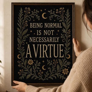 Op de afbeelding: Een rechthoekig ingelijst wandkunstwerk van zwarte stof met goudkleurig geborduurde tekst die luidt BEING NORMAL IS NOT NECESSARILY A VIRTUE. Het ontwerp bevat gouden bloemranken, bladeren en halve maan motieven rondom de belettering.