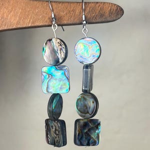 May include: A pair of dangle earrings featuring alternating circular and square abalone shell beads. Each earring displays iridescent blue, green, and purple hues with natural shell patterns. The beads are connected by silver-toned metal wire hooks.