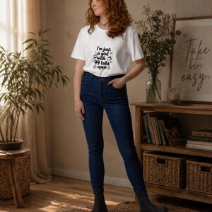 Op de afbeelding: Een wit T-shirt met korte mouwen en de zwarte tekst I'm just a girl with 99 tabs open, vergezeld van kleine iconen van een laptop, een koffiemok en een document, gecombineerd met een donkerblauwe spijkerbroek en zwarte enkellaarsjes.