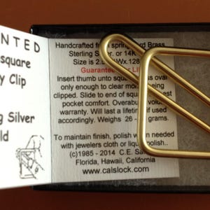 May include: A gold-coloured metal Thumbsquare money clip resting in a black box. The packaging includes text: PATENTED Thumbsquare Money Clip, Brass, Sterling Silver, 14K Gold, Personally Handcrafted in the U.S.A. since 1985, and calslock.com.