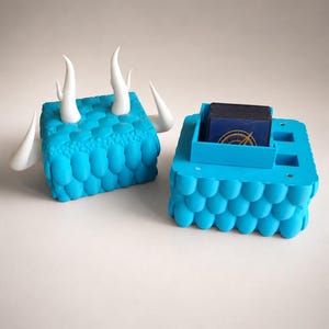 Puede incluir: Una caja rectangular de color azul brillante para guardar cartas con una superficie texturizada similar a escamas. La tapa presenta cuatro protuberancias blancas curvas en forma de cuernos. La base contiene un compartimento central con cartas.