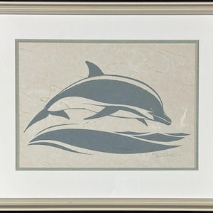 May include: A framed art print featuring a stylized gray dolphin leaping above a gray wave on a textured beige background. The artwork is enclosed in a light-colored wooden frame with a white mat border and a thin gray inner accent frame.