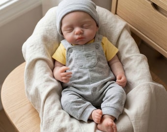 May include: A realistic baby doll with closed eyes, wearing a light grey beanie, a yellow short-sleeved shirt, and grey overalls featuring a subtle elephant pattern. The doll has a soft, lifelike appearance and is positioned in a seated posture.