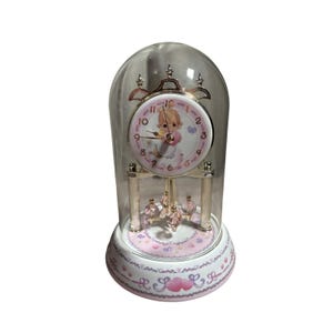 May include: A dome-shaped glass clock featuring a white face with pink numbers and a central illustration of a child holding a heart. The clock has gold-tone accents, a rotating pendulum with pink crystal-like beads, and a white base with pink hearts.