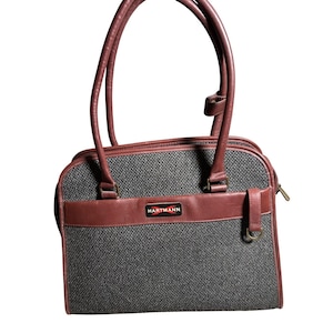 May include: A structured handbag featuring a grey textured fabric body with dark maroon leather trim and handles. A rectangular black label with the white text HARTMANN and a red graphic element is centered on the front leather panel.