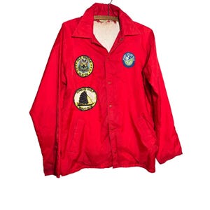 May include: A bright red windbreaker jacket featuring a pointed collar, long sleeves, and a snap-button front closure. The chest displays three embroidered patches: USS Reeves DLG-24, Tonkin Gulf Yacht Club, and a circular blue and white logo.