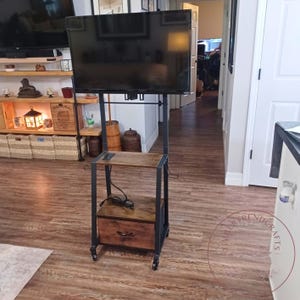 May include: A mobile television stand featuring a black metal frame with a top mount for a flat screen, a middle shelf with a power outlet, and a bottom storage drawer with a dark wood grain finish, all supported by four small caster wheels.
